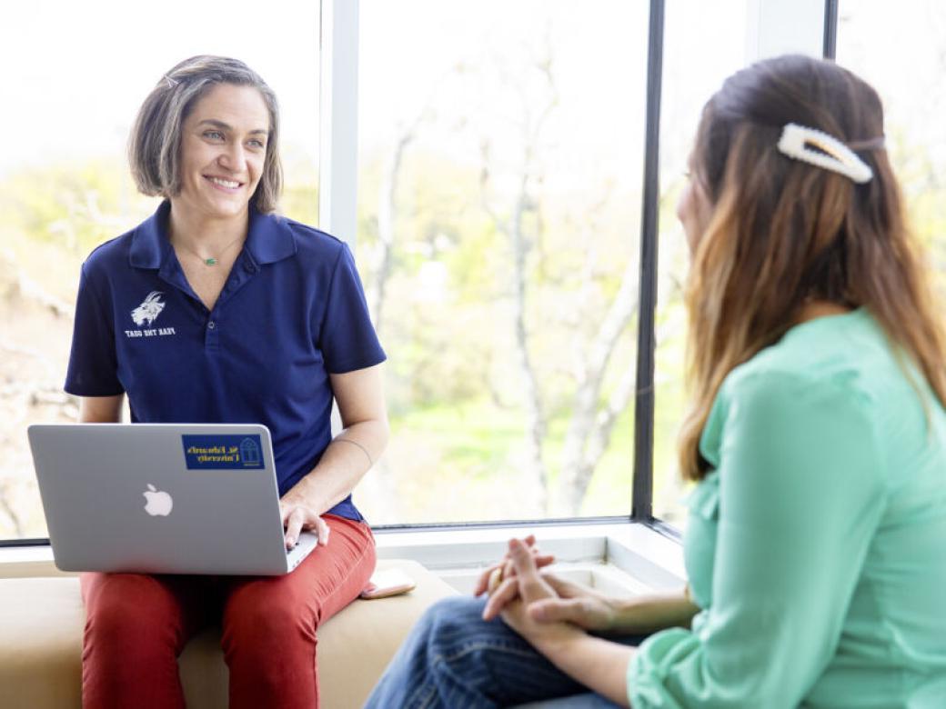 Graduate students sit on benches by a window and talk. One student is wearing a St. Edward's polo and has a laptop in their lap.