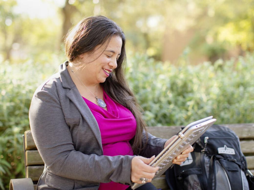 A graduate student sits on a bench with their backpack next to them holding a laptop in a tablet orientation. 