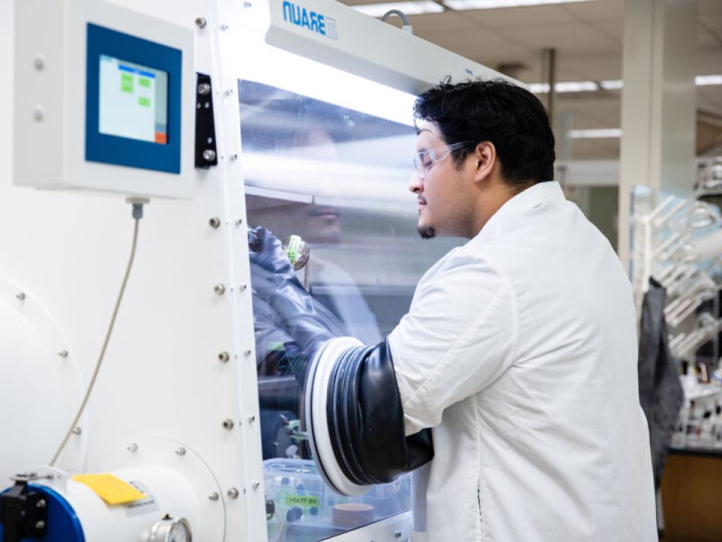 A student wearing a lab coat and protective glasses looks at bottles of chemicals using specialized equipment.