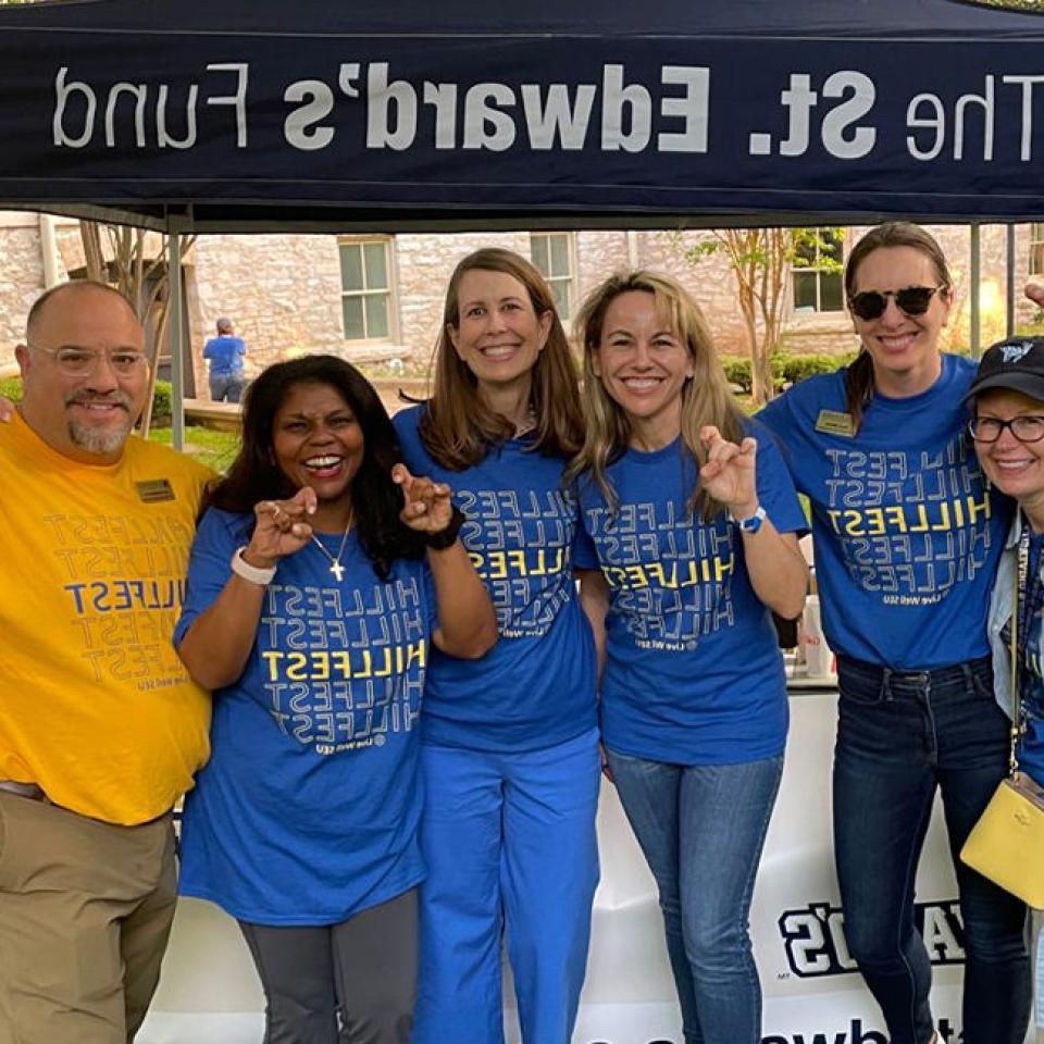 The image features six people standing together under a canopy that reads "The St. 爱德华的基金." 他们都在微笑 and making hand gestures, likely the university's hand sign. Most of them are wearing blue T-shirts with "HILLFEST" printed on them, 说明他们是同一事件的一部分. 一个人穿着黄色版的t恤. 背景是校园的建筑和绿化, 说明这是一场大学活动.