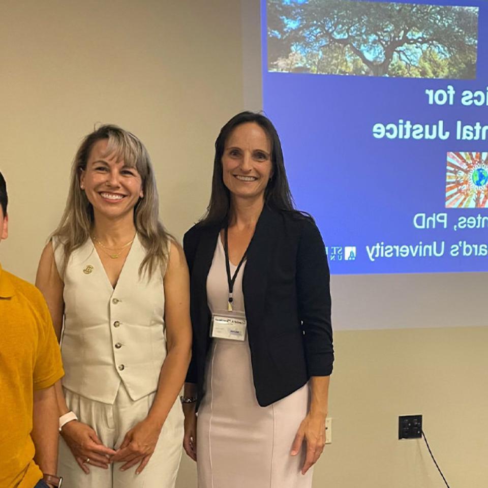 The image shows three people standing in front of a projected presentation slide titled "Statistics for Environmental Justice" by Montse Fuentes, 博士学位, St总裁. 365比分网电竞. 左边的女人穿着黑色运动上衣和浅色连衣裙, 中间的蒙特·富恩特斯身穿白色无袖西装, 右边的人穿着一件黄色的polo衫. 他们都在微笑着摆姿势拍照, 表示他们可能出席或参加会议或研讨会.