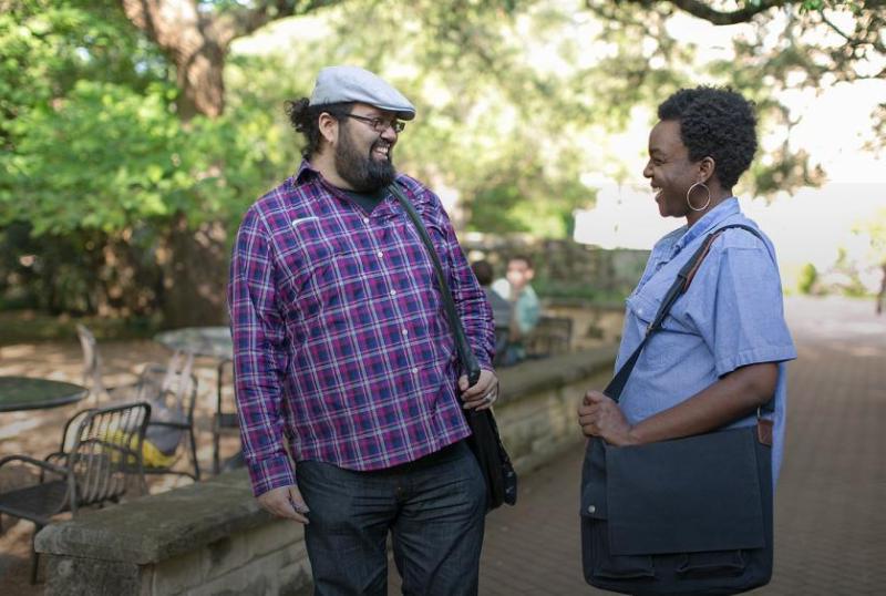 The image shows two people standing outdoors, facing each other. The person on the left is wearing a short-sleeved shirt with vertical stripes, dark pants, and is carrying a shoulder bag. The person on the right is dressed in a plaid shirt with long sleeves, jeans, and a white cap. In the background, there's a natural setting with trees and what appears to be a park area. You can see stone edging and outdoor furniture, and there are other people further in the background walking away from the camera.