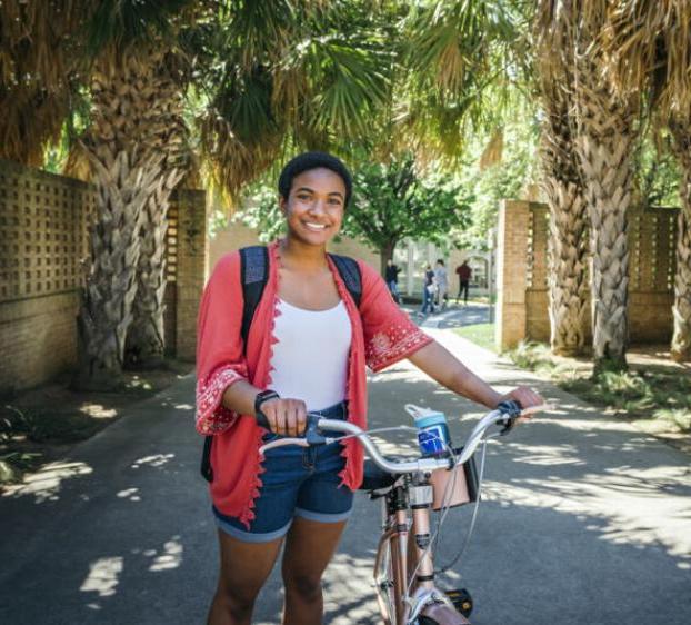 This image shows a young woman with a joyful expression, riding a bicycle through a sunny, tree-lined pathway. She wears a white tank top, red cardigan, and denim shorts, complemented by a backpack. Her bike is equipped with a small front basket containing a blue item.